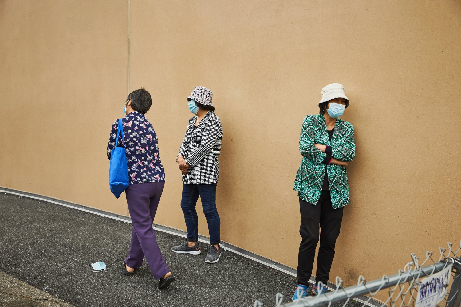 Three women in face masks stand against a beige wall, two with patterned jackets, one carrying a blue tote bag, in an outdoor urban setting.