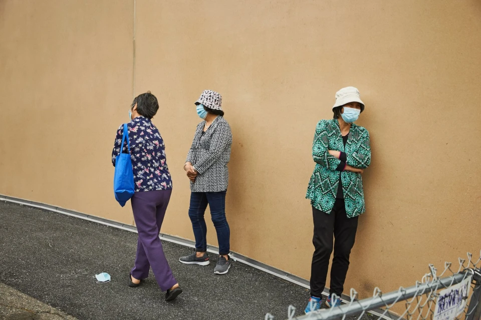 Three women in face masks stand against a beige wall, two with patterned jackets, one carrying a blue tote bag, in an outdoor urban setting.