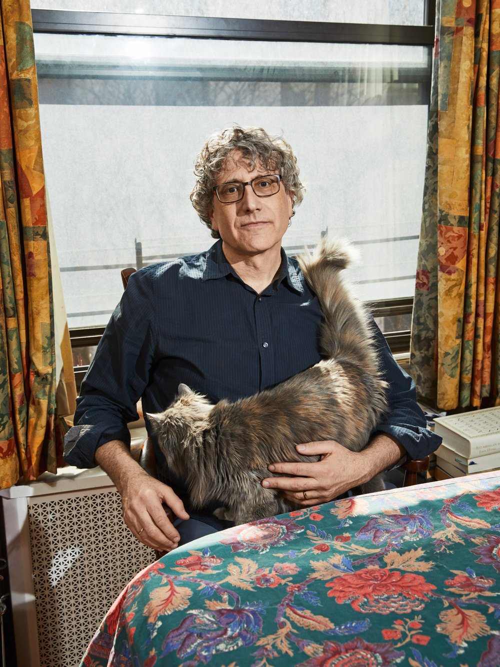 Indoor portrait of a man with curly hair and glasses holding a grey cat, with a floral tablecloth, radiator, and curtains in view.