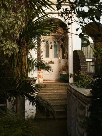 A white house entrance with an arched doorway, concrete steps, and tropical plants surrounding the scene in a residential area.