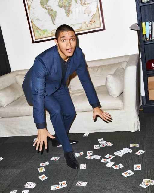 Trevor Noah poses indoors wearing a blue suit, looking surprised on a floor with scattered playing cards, a white couch behind him.
