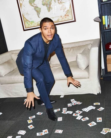Trevor Noah poses indoors wearing a blue suit, looking surprised on a floor with scattered playing cards, a white couch behind him.