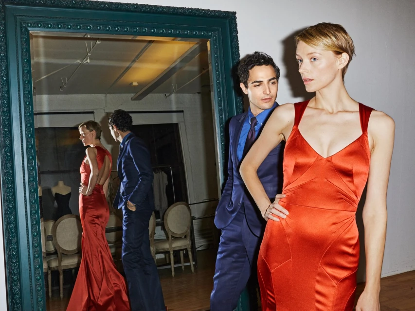 Fashionable adult female in a red gown poses next to an adult male in a blue suit in an indoor setting with an ornate mirror.