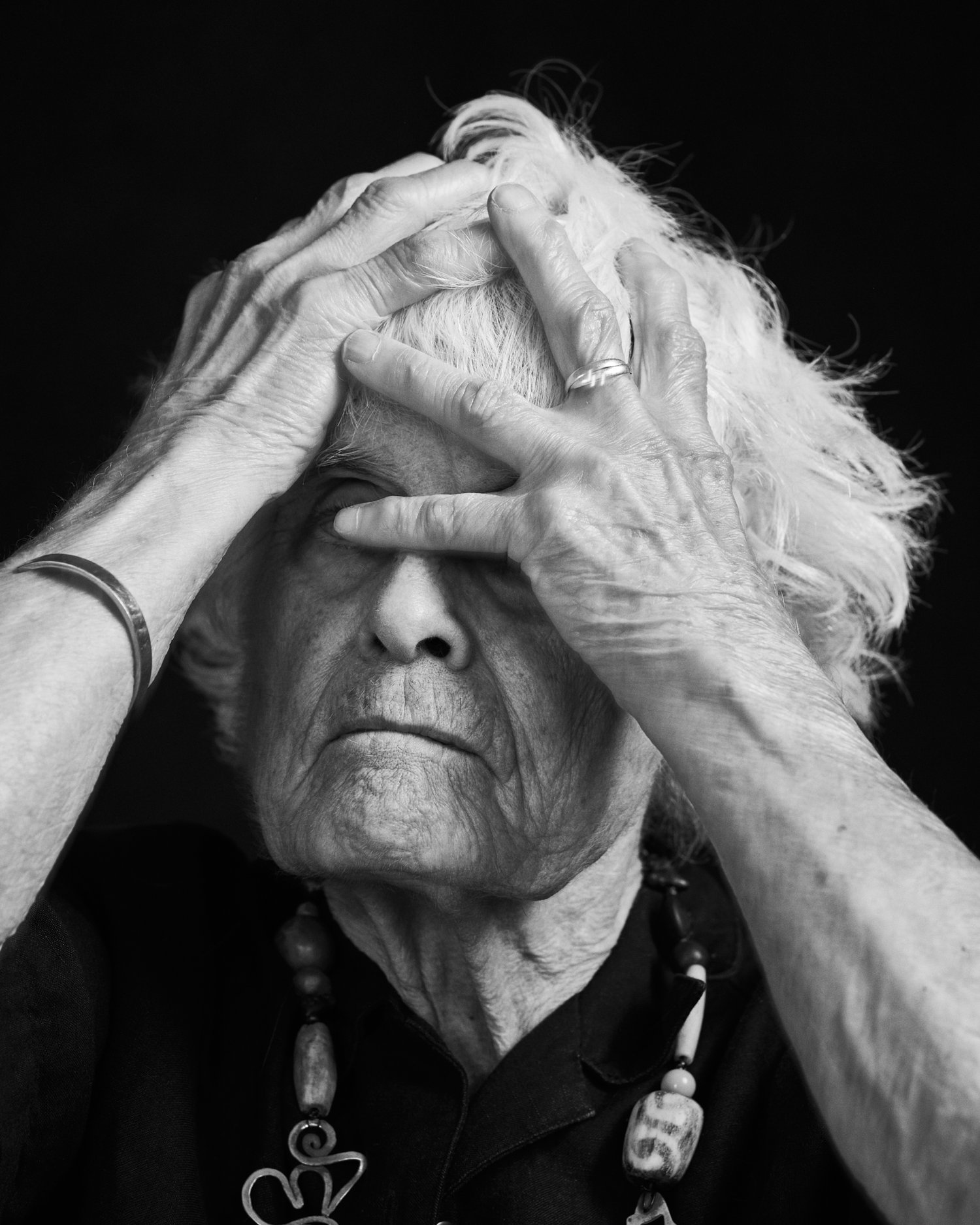 Elderly person in black and white portrait, their hands covering their forehead, wearing a beaded necklace and dark collared shirt.