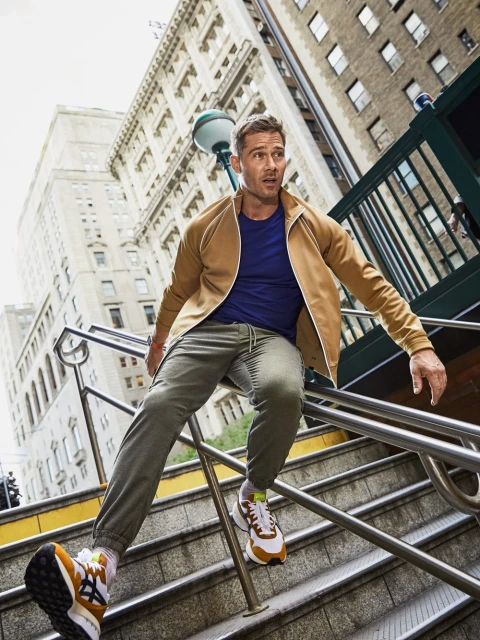 Stylish man in a tan jacket and blue t-shirt descends an urban staircase with a tall building in the background on a sunny day.