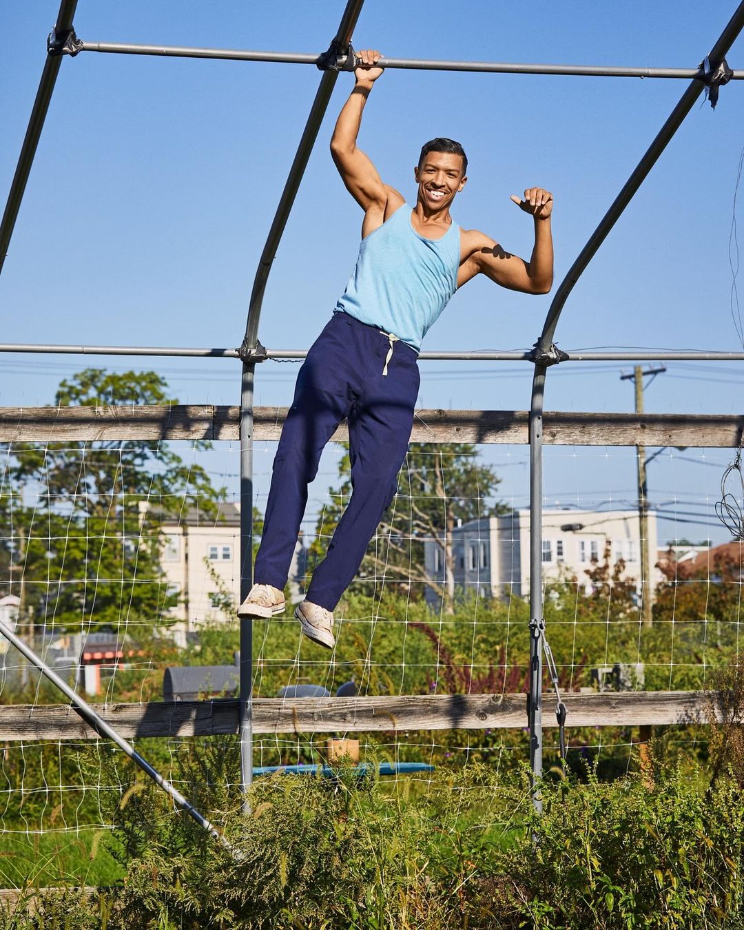Athletic man hanging from metal bars outdoors on a sunny day. Wearing a light blue tank top and dark pants, he is smiling and looks strong.