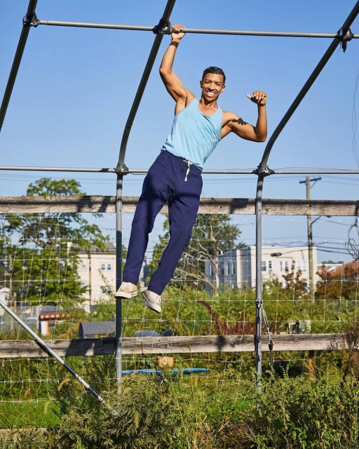 Athletic man hanging from metal bars outdoors on a sunny day. Wearing a light blue tank top and dark pants, he is smiling and looks strong.