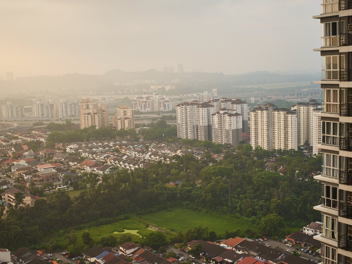 Wide outdoor shot of a dense urban landscape with residential buildings, high-rise apartments, and lush green trees under a hazy sky.