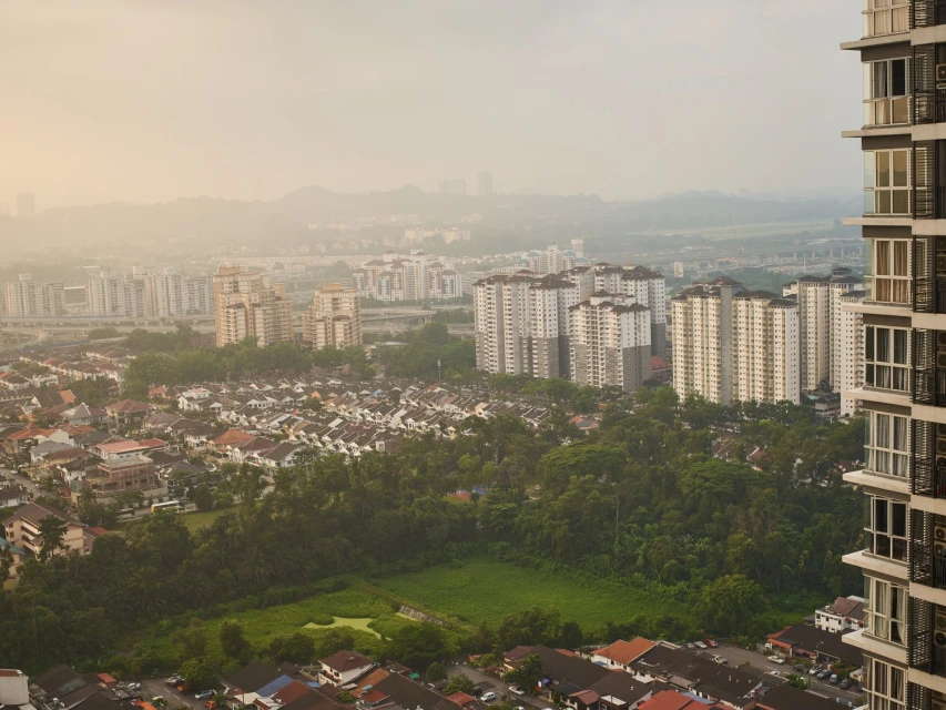 Wide outdoor shot of a dense urban landscape with residential buildings, high-rise apartments, and lush green trees under a hazy sky.