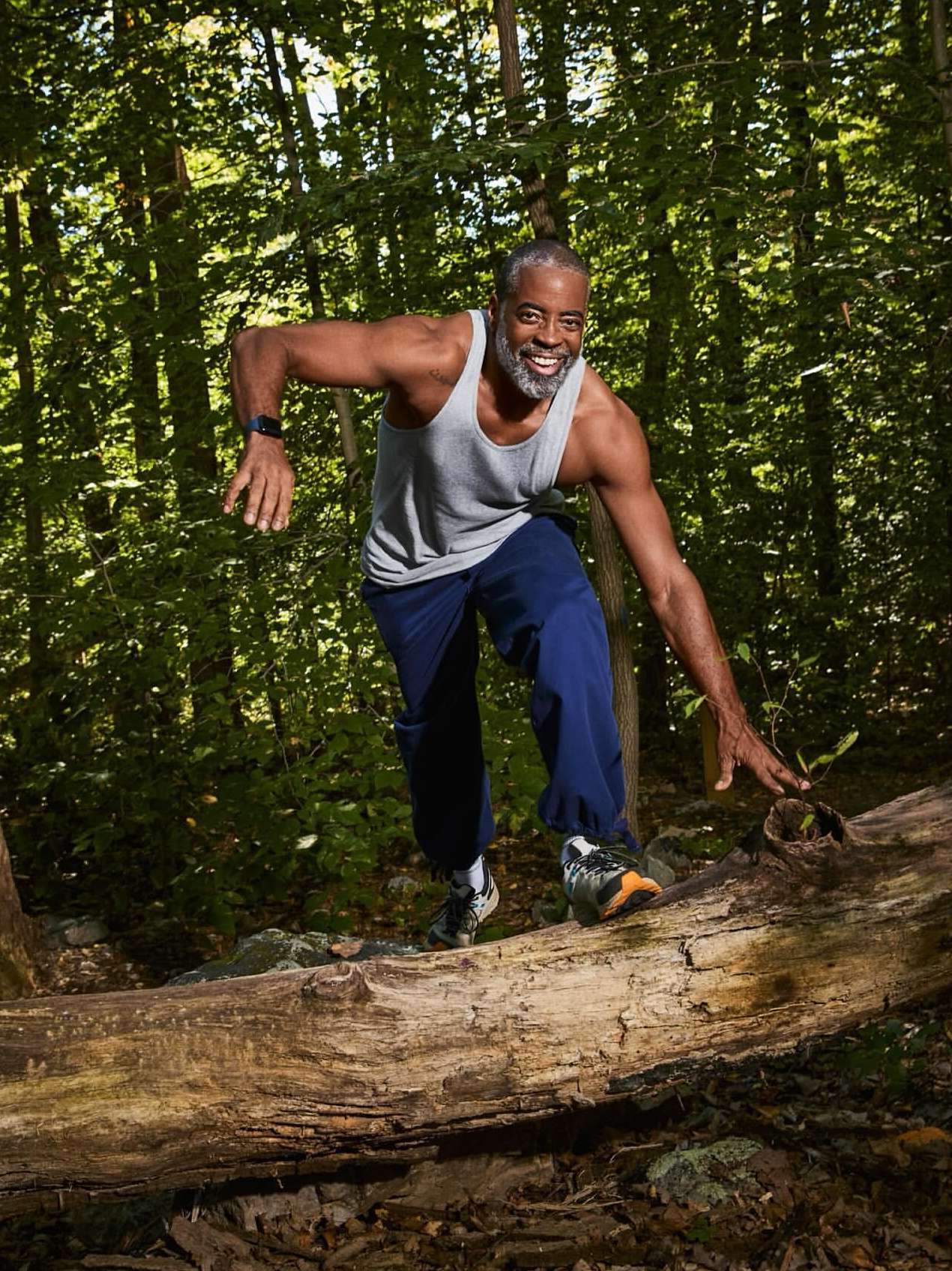 An African-American man with a gray beard crosses over a fallen tree trunk in a forest, wearing a tank top and workout pants.