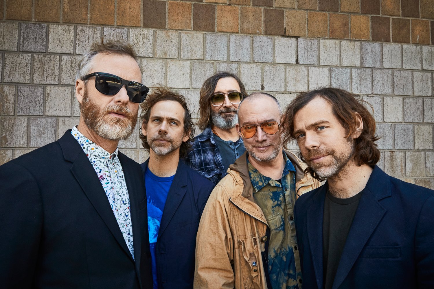 Group portrait of five adult men with beards in casual and semi-formal outfits posing against a textured beige brick wall outdoors.