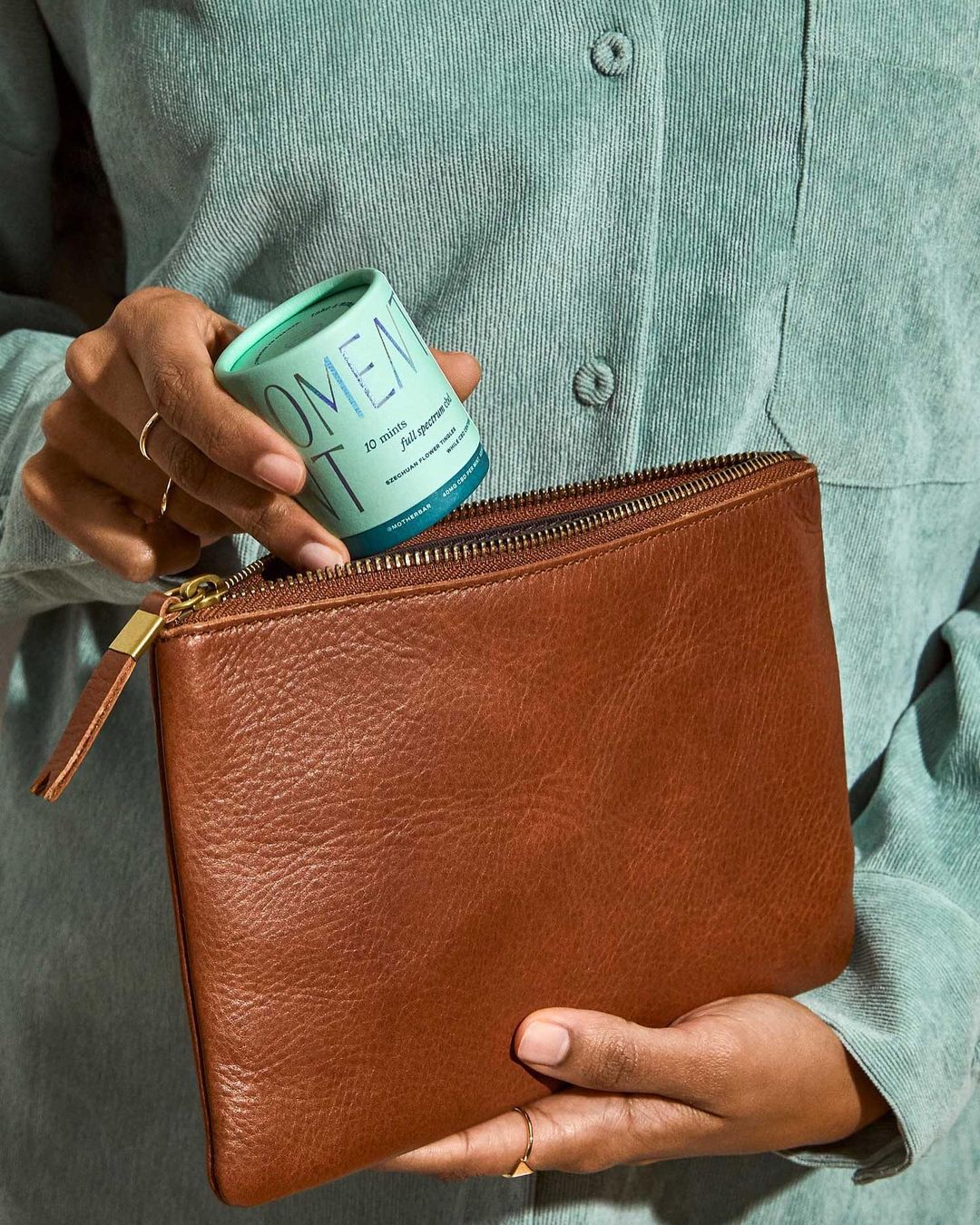 A woman in a blue shirt is holding a brown leather clutch and a turquoise container in an indoor setting. The shot is a medium close up.