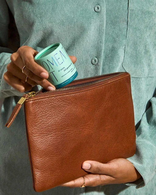 A woman in a blue shirt is holding a brown leather clutch and a turquoise container in an indoor setting. The shot is a medium close up.