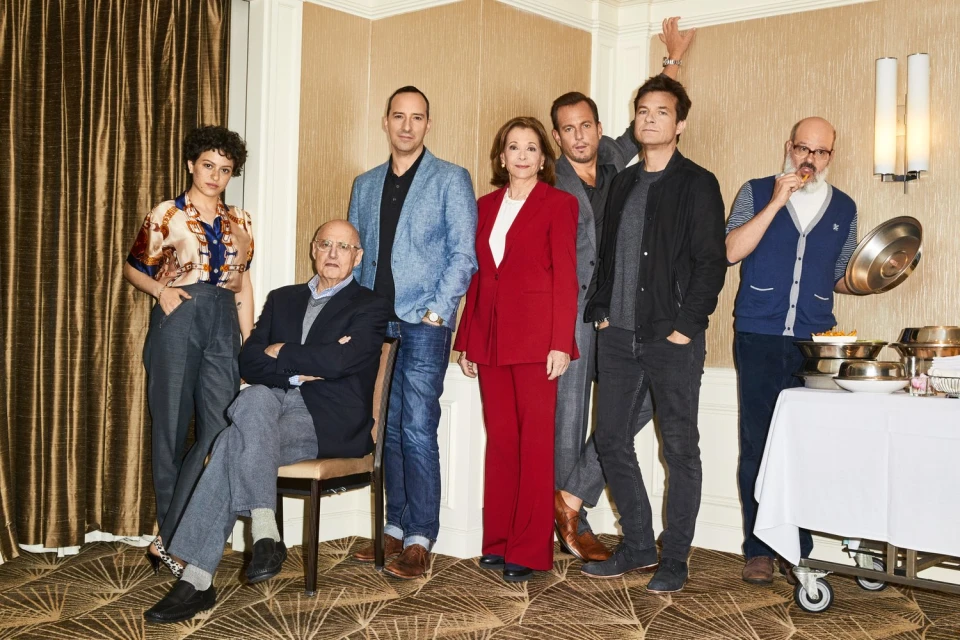 Group portrait of seven people in a room, including one seated elderly man, one woman in a red suit, and a man with a buffet server.