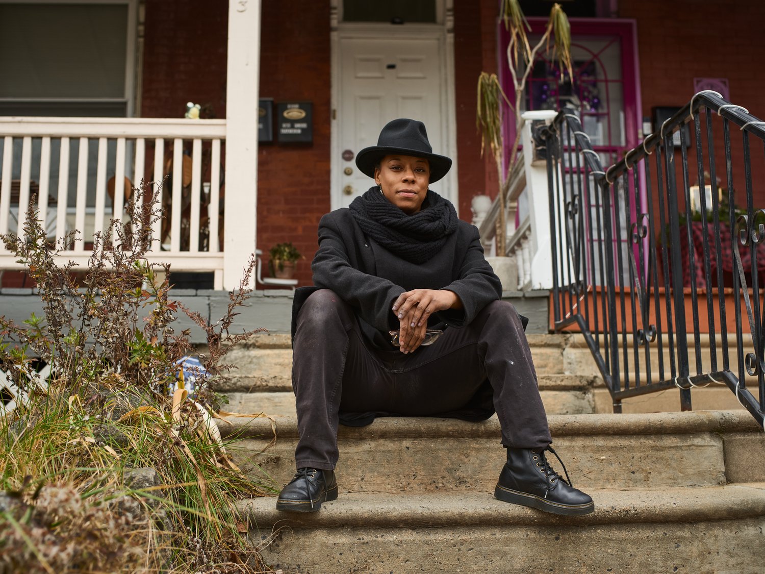Person sits on porch steps wearing black hat, scarf, coat, pants, and boots in an outdoor urban environment with house in background.