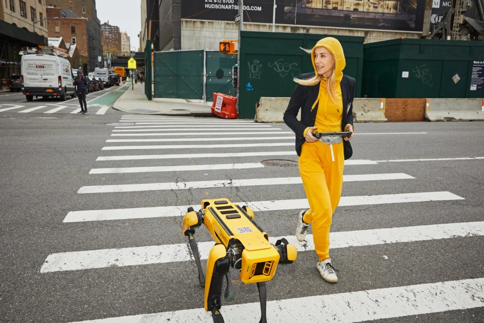 Woman runs across crosswalk with yellow robotic dog in an urban setting, wearing yellow tracksuit and black blazer.