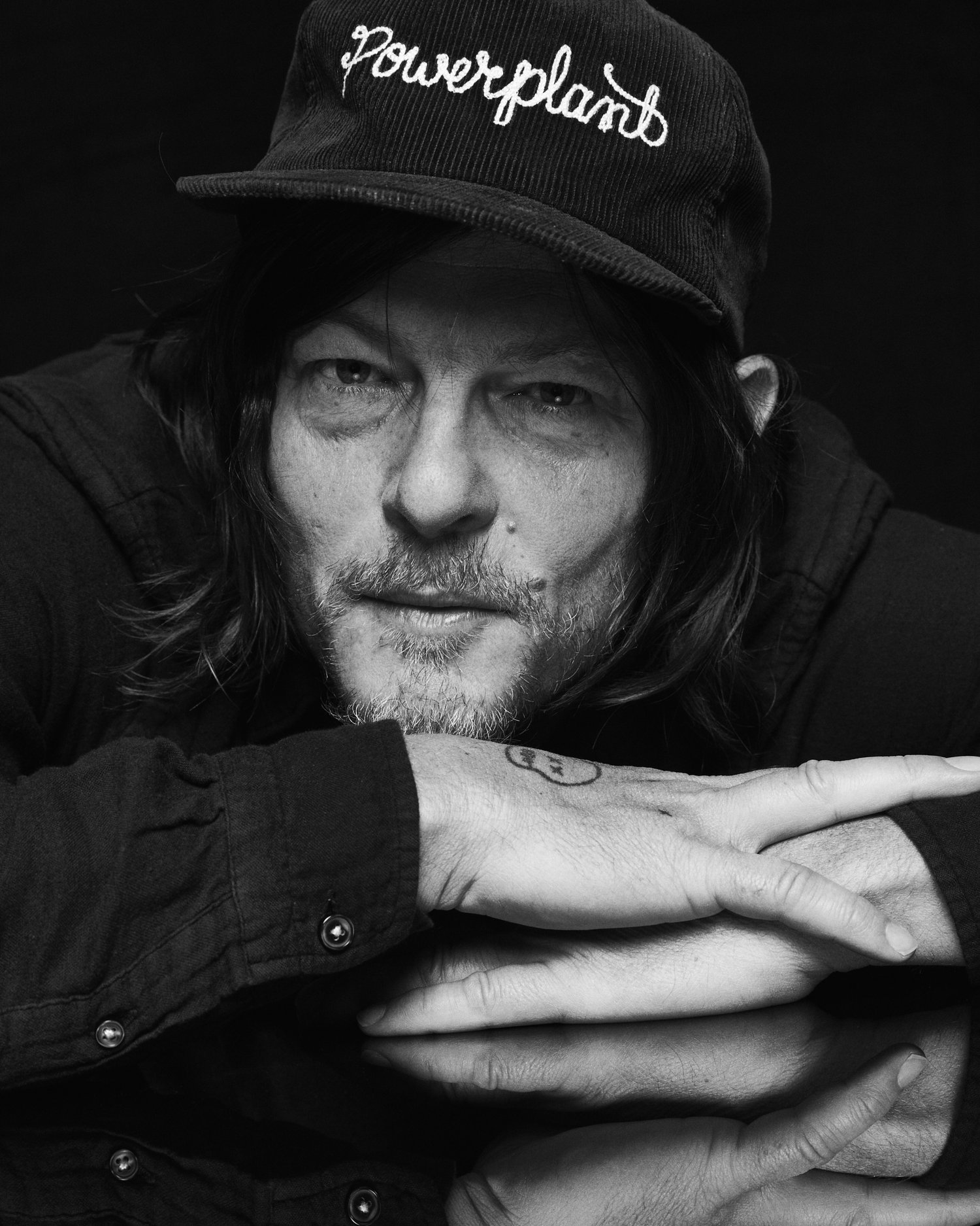 Black and white portrait of adult male with cap and long hair resting head on crossed arms on black reflective surface, looking at camera.