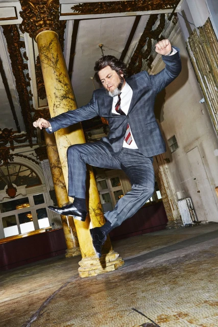 Adult male wearing a grey suit, white shirt and striped tie jumping in the air in a room with pillars and ornate details.