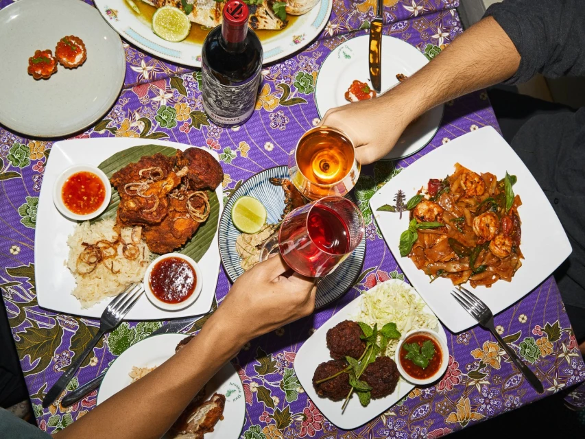 Overhead view of people toasting wine glasses over a table laden with various dishes, including fried chicken, noodles, and appetizers.