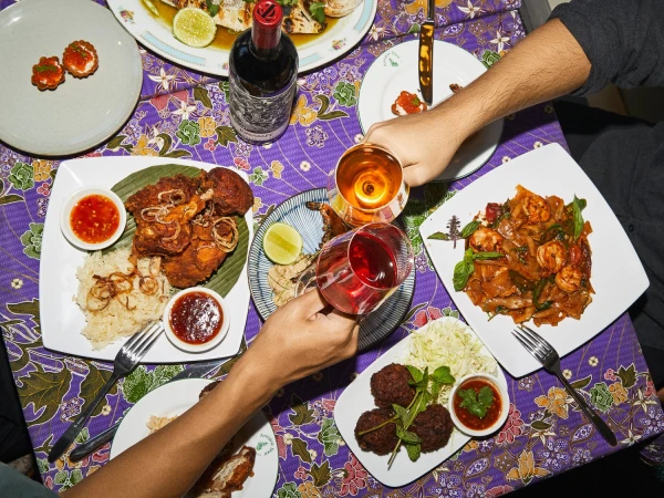 Overhead view of people toasting wine glasses over a table laden with various dishes, including fried chicken, noodles, and appetizers.