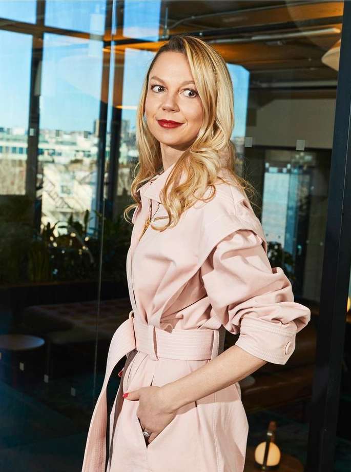 Woman in a pink dress poses indoors near a window, showcasing her blonde hair and red lipstick, in a bright and stylish setting.