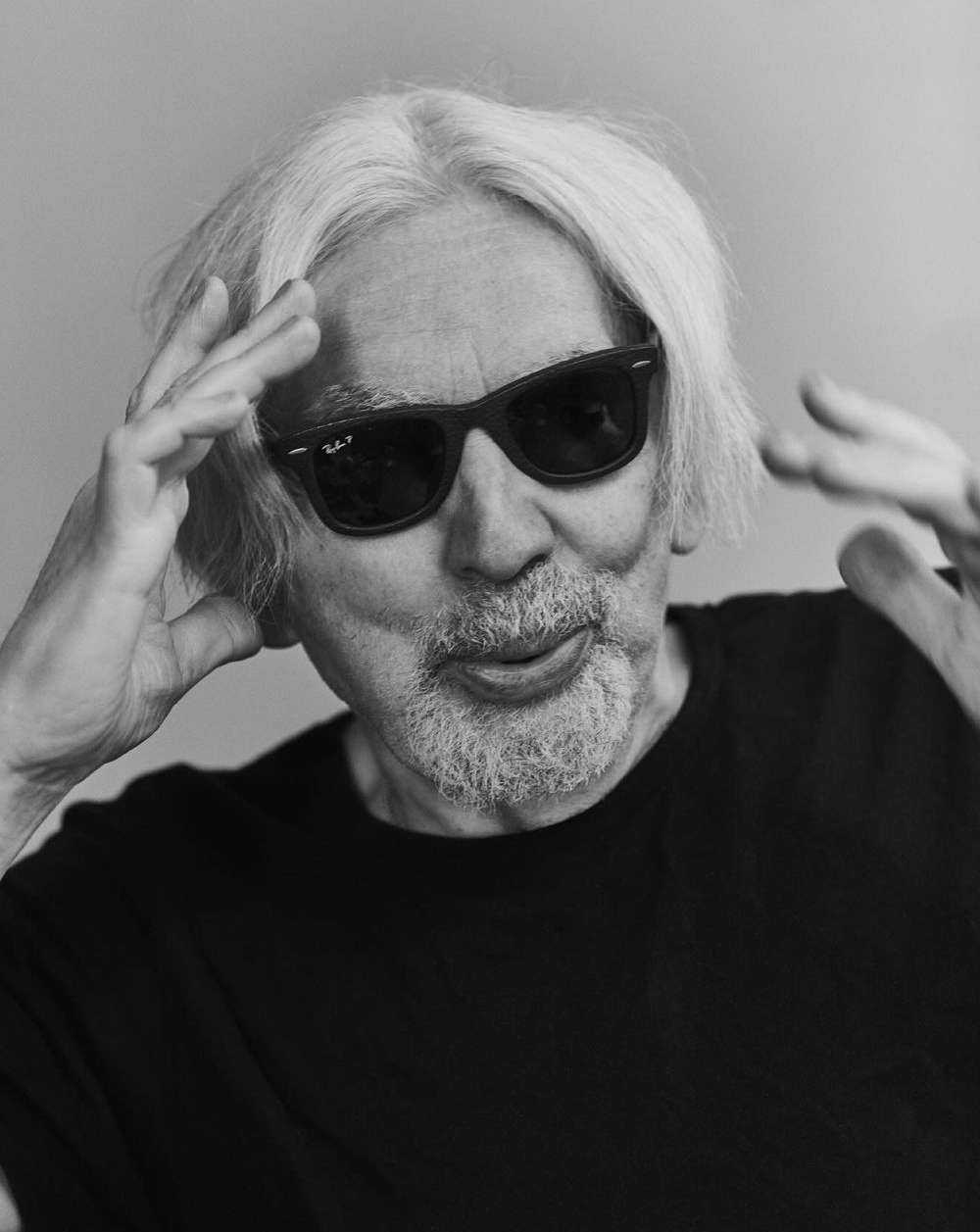 Black and white portrait of an elderly man wearing dark glasses, sticking his tongue out, and holding his hands up to his head.