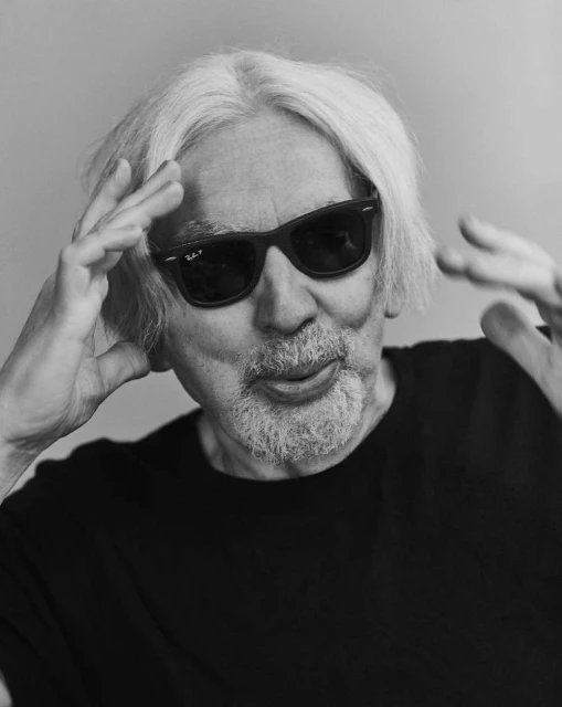 Black and white portrait of an elderly man wearing dark glasses, sticking his tongue out, and holding his hands up to his head.