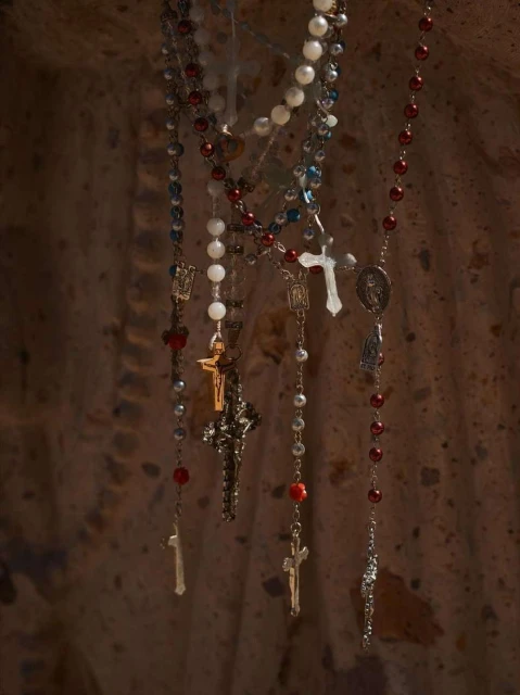 Several rosaries with cross pendants are hanging against a textured brown background in a studio setting. Beads and metal chains visible.