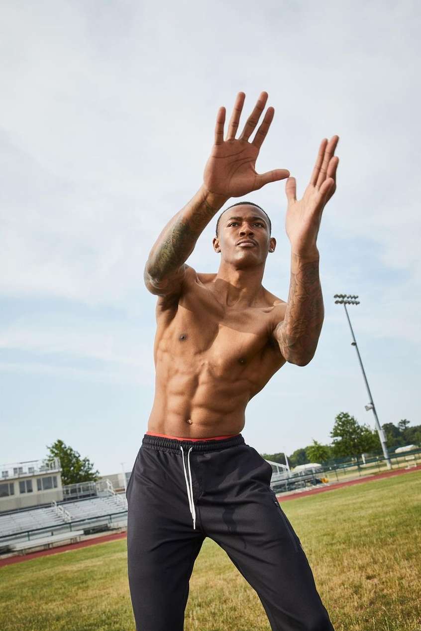 Athletic African-American man with bare torso and arms raised on a sports field under a bright sky, captured from a low angle.