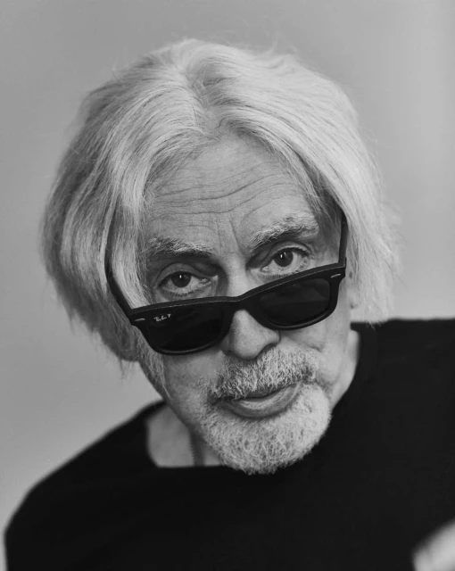 Black and white portrait of an older man with grey hair and a goatee, wearing dark sunglasses and a black shirt. Indoors.