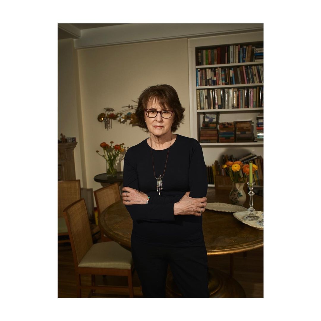 Elderly woman wearing dark long-sleeved shirt and glasses with arms crossed in a living room interior by Aaron Richter