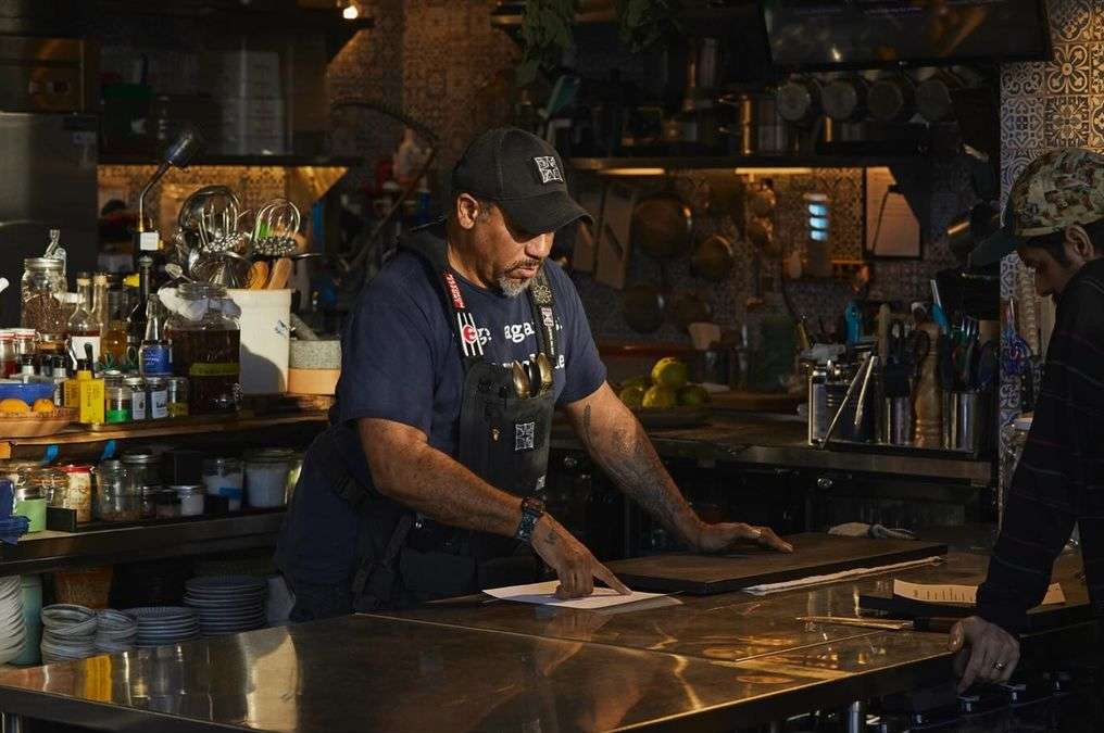Two chefs review a document at a stainless steel kitchen counter in a warm-lit, restaurant kitchen with cookware and tiled wall.