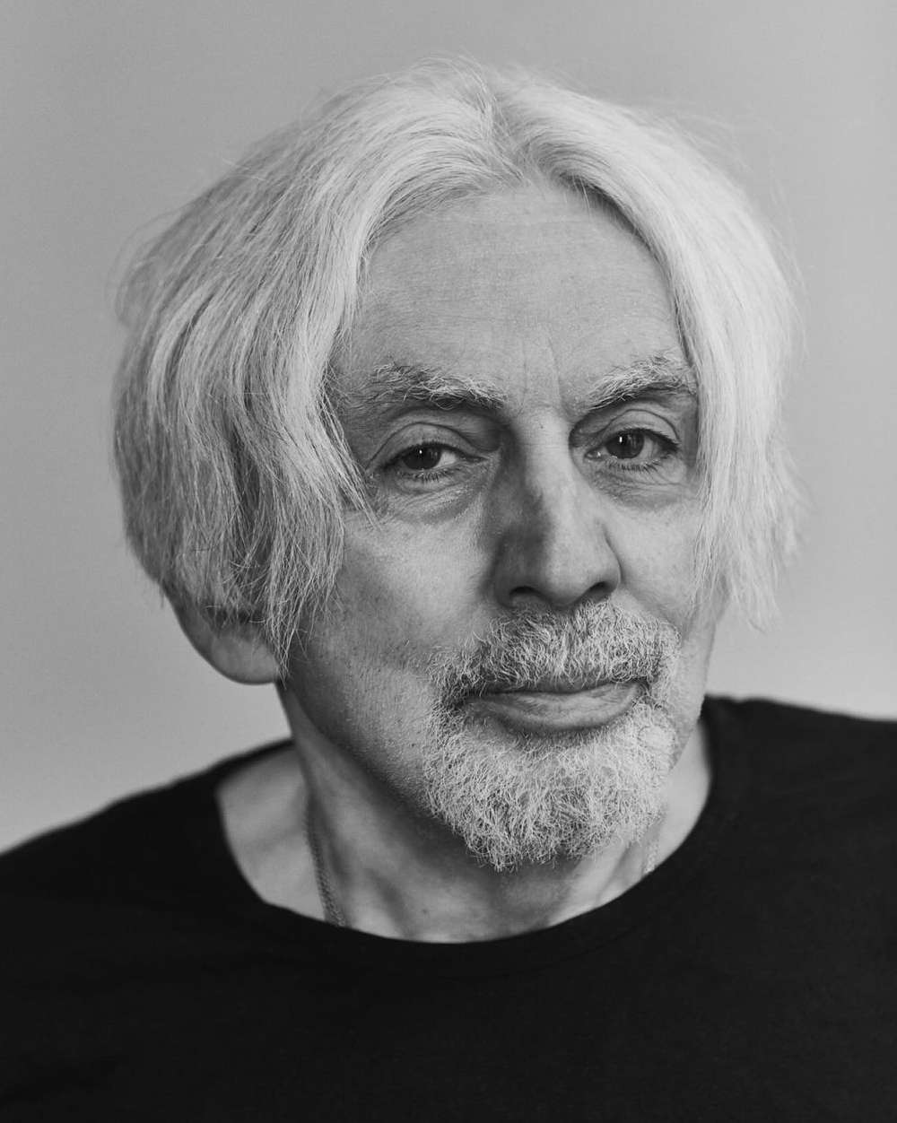 Black and white portrait of an elderly man with long white hair and a white beard, wearing a black shirt against a gray background.