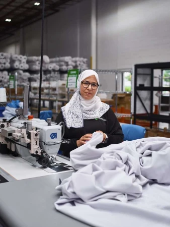 Adult woman in hijab working at sewing machine in factory, producing textile product with large rolls of material in background.