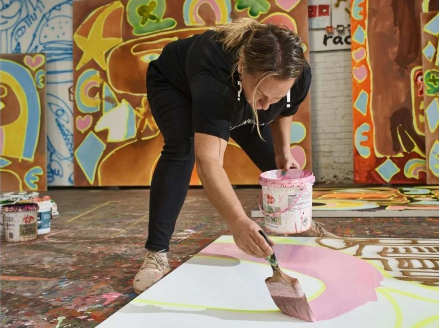 Female artist creating colorful paintings in a studio, using a large paintbrush on a large canvas with other canvases around.