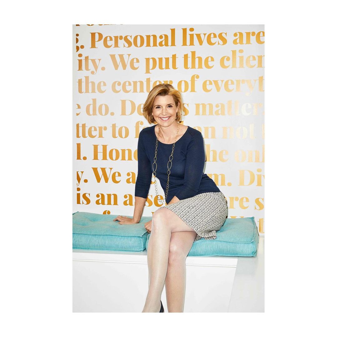 A smiling middle-aged woman wearing a navy blue top and patterned skirt seated on a turquoise cushion against a text-covered wall