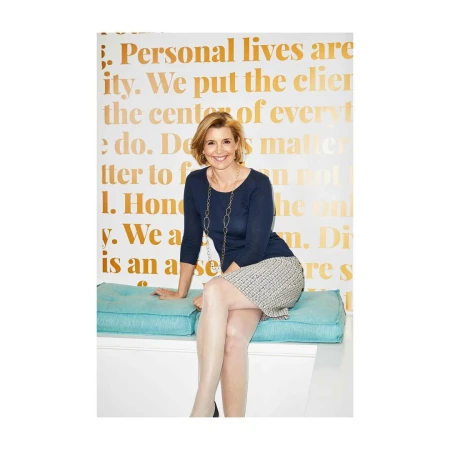 A smiling middle-aged woman wearing a navy blue top and patterned skirt seated on a turquoise cushion against a text-covered wall