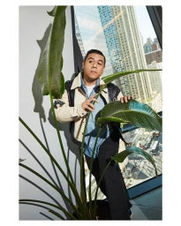 Man standing indoors next to a large potted plant near a tall glass window overlooking a city skyline.