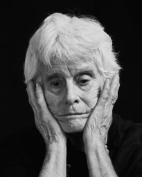 Portrait of an elderly woman with short white hair holding her face in her hands against a solid black background.