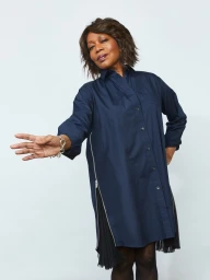 Older woman with short dark hair wearing a navy blue shirt dress extending hand forward in studio
