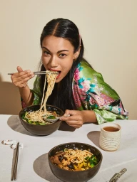 Woman in a green floral robe eating noodles with chopsticks from a dark ceramic bowl at a table.