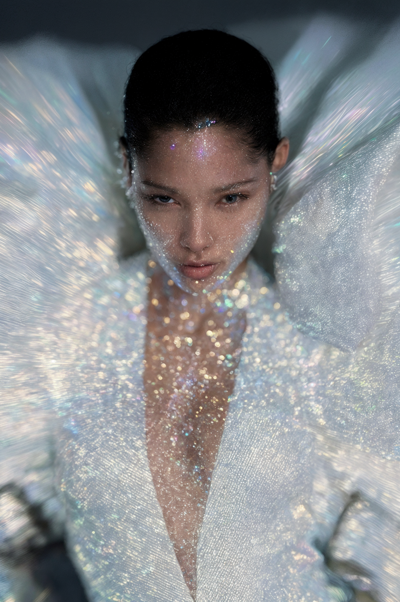 Portrait of a young woman with glitter on her face and wearing an iridescent white jacket, gazing directly at the camera in a studio setting.