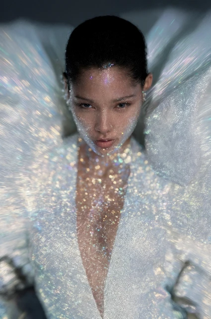 Portrait of a young woman with glitter on her face and wearing an iridescent white jacket, gazing directly at the camera in a studio setting.