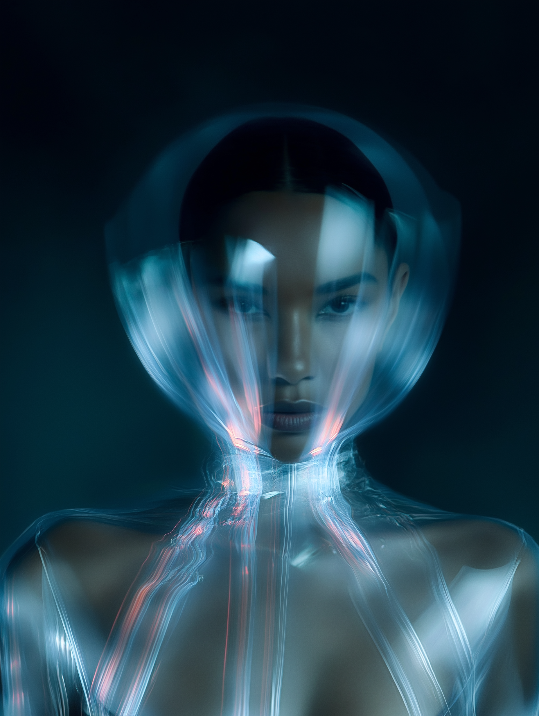 Dramatic studio portrait of an adult female wearing a futuristic clear plastic hood and garment with red accents against a teal background.