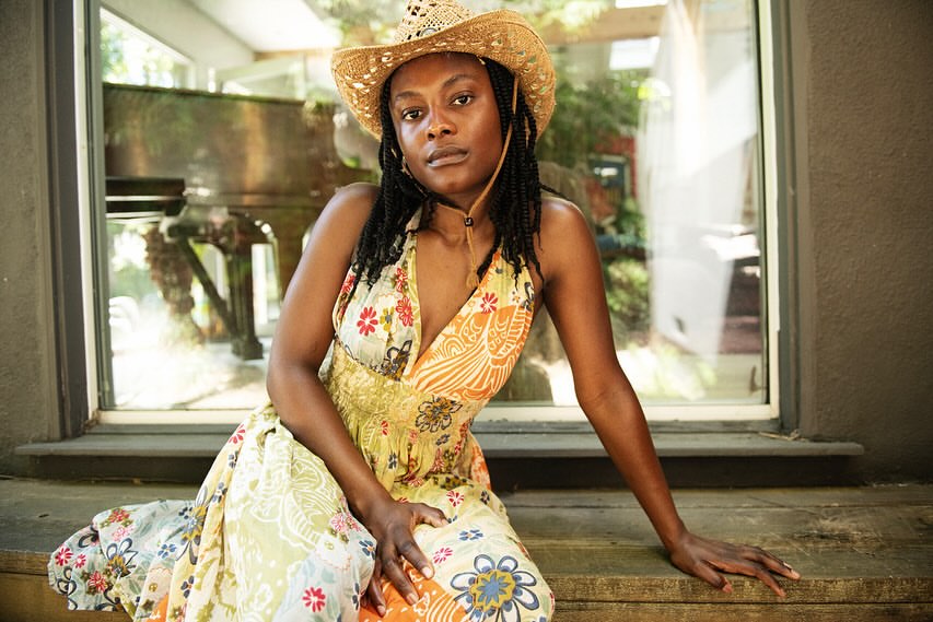 A woman with braided hair wears a straw cowboy hat and floral dress while posing in front of a window with plants and a piano visible.