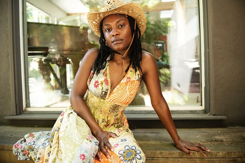 A woman with braided hair wears a straw cowboy hat and floral dress while posing in front of a window with plants and a piano visible.