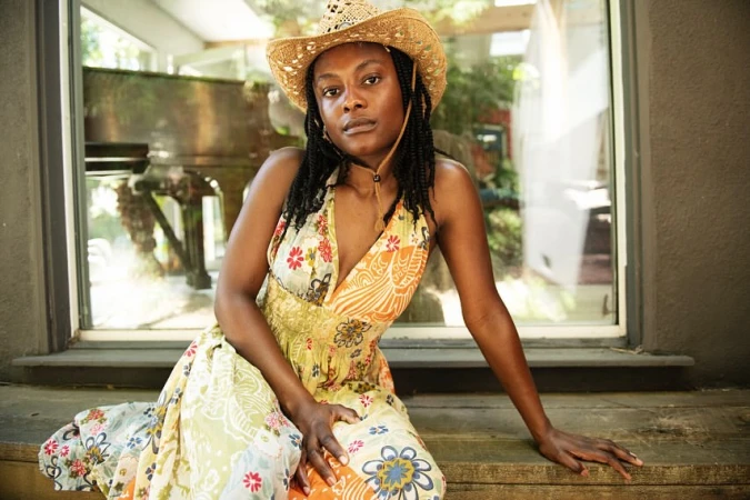 A woman with braided hair wears a straw cowboy hat and floral dress while posing in front of a window with plants and a piano visible.