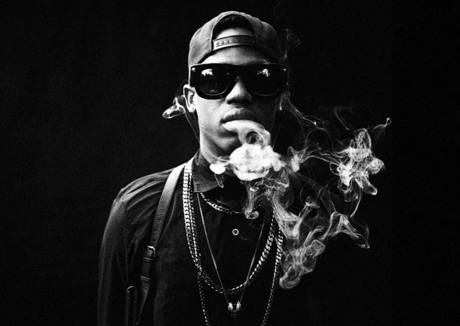 Black and white portrait of a stylish man in sunglasses and a cap, exhaling a cloud of smoke against a dark backdrop, with chain necklaces.