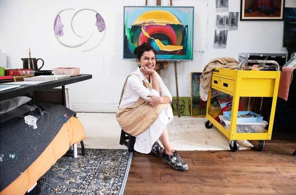 An artist sits in her studio, smiling, surrounded by her colorful paintings, art supplies, and personal touches in a creative indoor space.