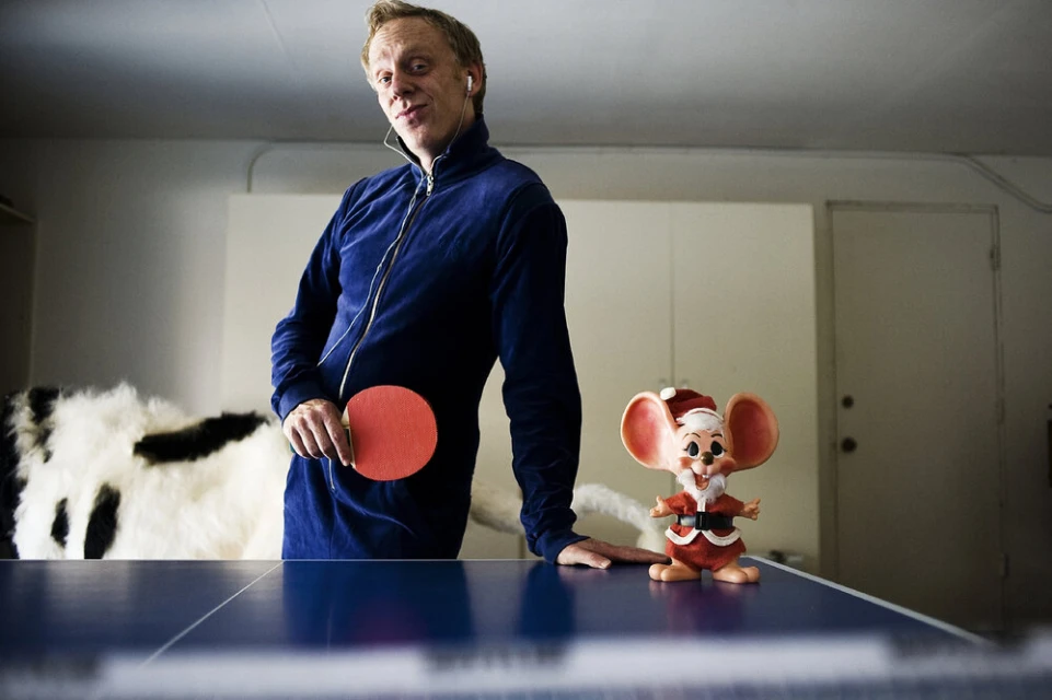 Man in blue tracksuit holding ping pong paddle standing next to a Christmas mouse figurine on a blue ping pong table in an indoor setting.